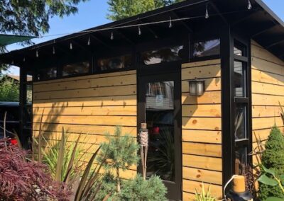 A modern wooden garden shed with black trim surrounded by lush greenery.