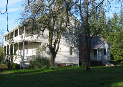 Two-story white house with a wraparound porch and a separate gazebo set amidst trees under a clear sky.