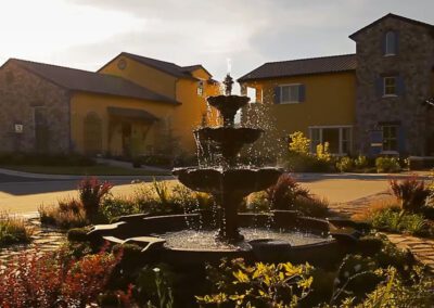 A fountain in the foreground with water cascading down multiple tiers, set against the backdrop of a sunny sky and stone buildings with yellow accents.