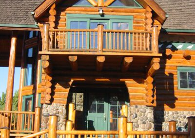A log cabin house with a stone foundation and a wooden balcony under a clear blue sky.
