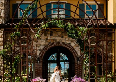 Bride walking through a garden archway in front of an elegant stone building.