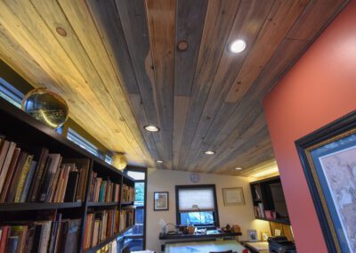 Interior view of a room with a wooden ceiling, recessed lighting, a bookshelf, and a desk by the window.