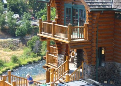A log cabin house by a river with a person and a vehicle in the foreground.