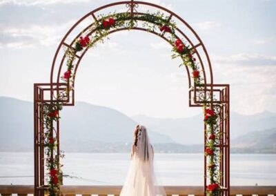 Bride standing under a floral archway with scenic lake and mountains in the background.