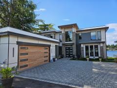 Modern two-story house with large windows, a flat roof, and a wooden double garage door, situated by a lake under a clear blue sky.