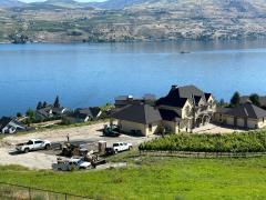 Large house under construction near a lake, with construction vehicles and materials in the driveway; hills and water are visible in the background.
