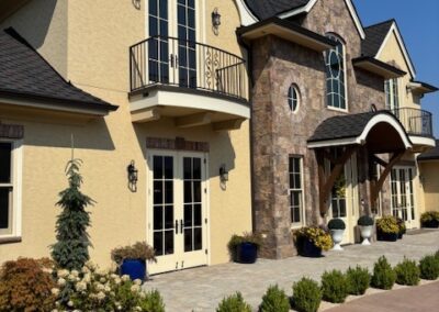 Two-story house with yellow stucco and stone exterior, arched windows, balcony, wooden front awning, and landscaped front yard.