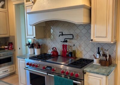 A modern kitchen with a large gas stove, double oven, red knobs, green countertop, wood cabinets, and utensils arranged beside the stove. A kettle and spices sit on the stove.