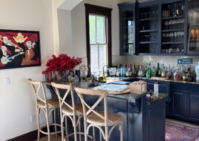 Home bar with wooden stools, navy cabinetry, glassware, and assorted bottles. A framed painting and vase with red flowers decorate the space. Natural light comes through a window.