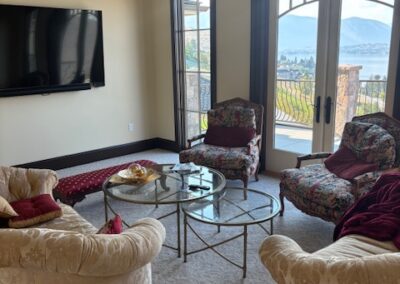 A living room with four armchairs around a glass coffee table, a wall-mounted TV, large windows, and a view of an outdoor balcony and mountains.