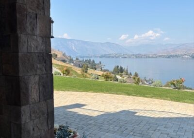 Stone archway frames a patio with pavers, overlooking a lake, distant hills, and mountains under a clear blue sky. A flower pot sits in the foreground.