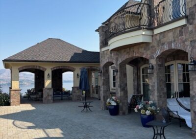 Stone house with arched patio, upper balcony, outdoor seating, potted plants, and mountain views in the background under a clear sky.