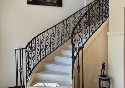 Curved staircase with ornate black railing, a framed black-and-white photo on the wall, and a small round table holding a sculpture at the base of the stairs.