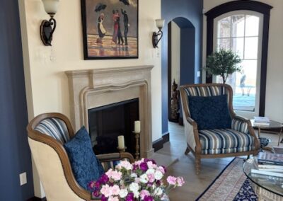 A living room with two armchairs by a fireplace, a floral arrangement on a small round table, and a painting above the mantel. A patterned rug and large window are also visible.